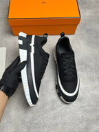 A pair of black low-top sneakers with chunky white and black soles, shown on a gray concrete floor with an orange shoebox in the background. One shoe is held by a gloved hand.