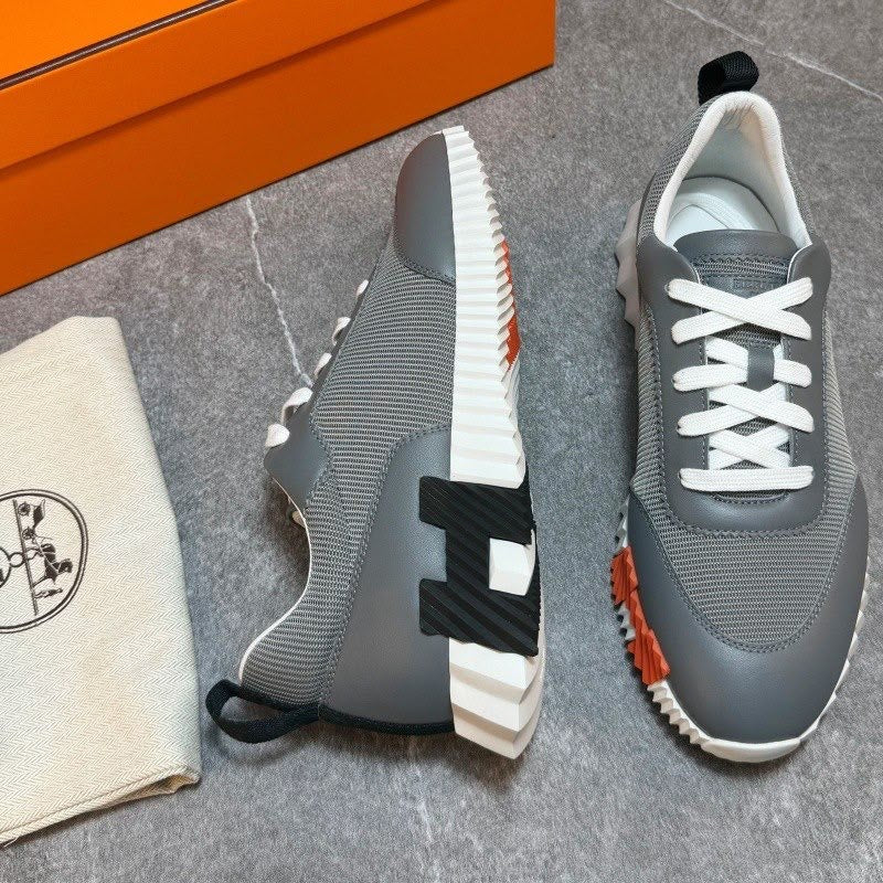A pair of gray knit sneakers with white laces, a thick white and black midsole, and orange accents, displayed next to an orange shoebox and a beige dust bag with a logo on a gray marble floor.