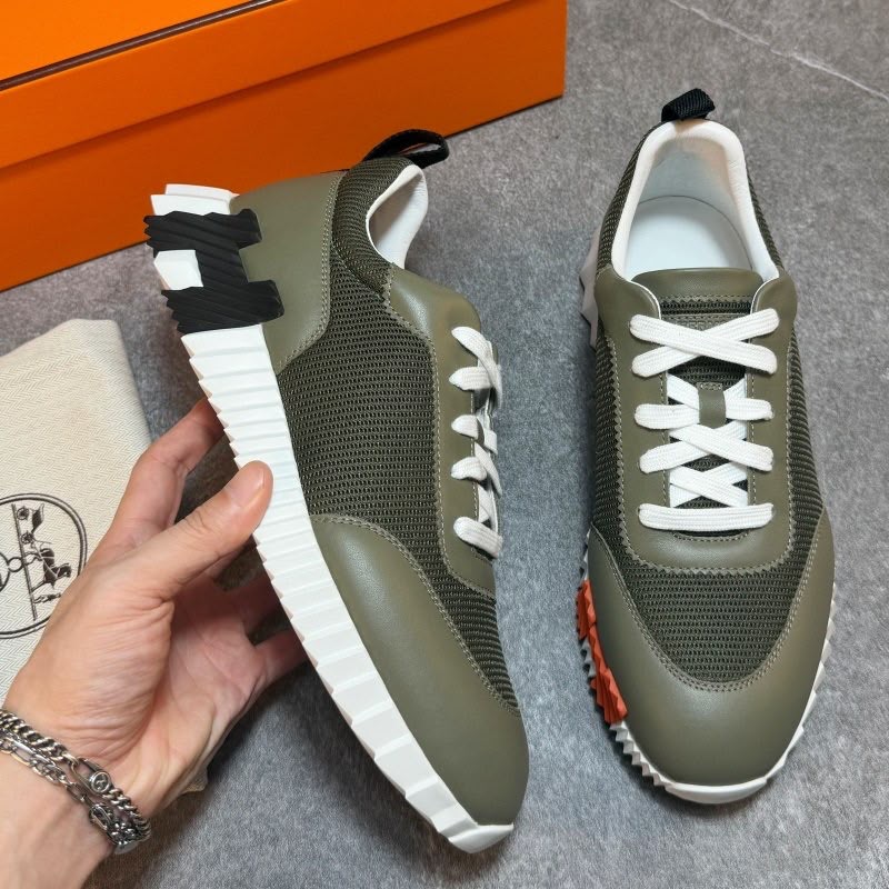 A pair of olive green and white low-top sneakers shown on a gray floor, with one shoe held by a hand and the other placed beside it. An orange box and a branded dust bag are partially visible in the background.