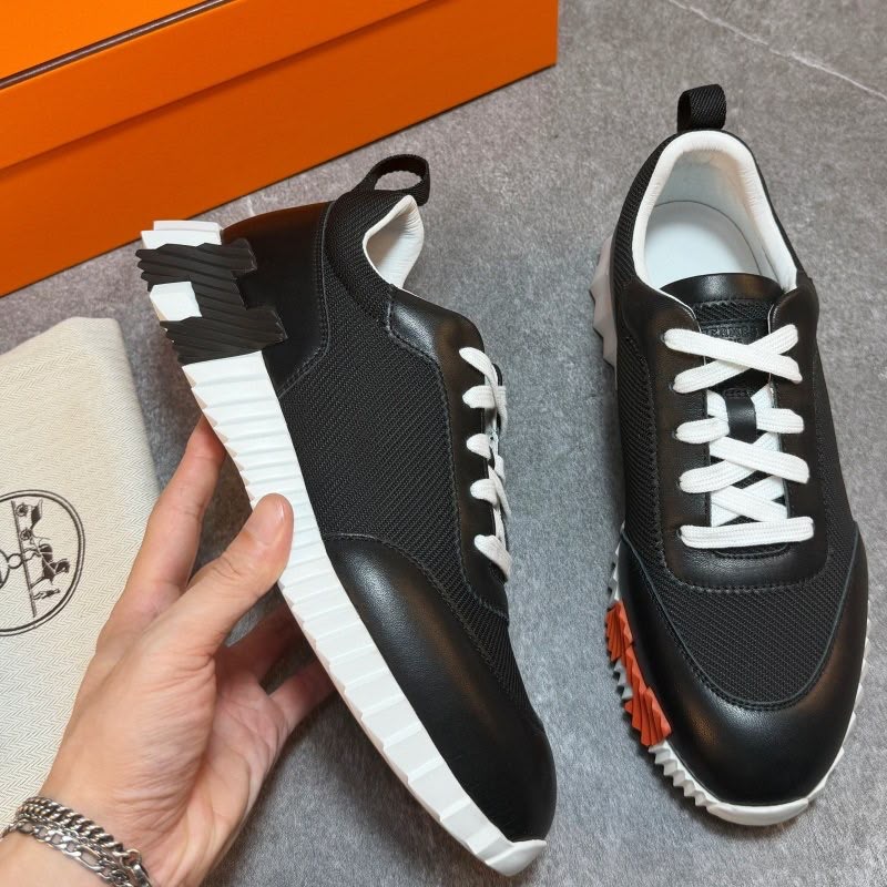 A pair of black low-top sneakers with white laces, white ridged soles, and orange accents, shown on a gray floor with an orange box and a cream-colored branded dust bag. A hand is holding one shoe.
