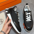 A pair of black low-top sneakers with white laces, white ridged soles, and orange accents, shown on a gray floor with an orange box and a cream-colored branded dust bag. A hand is holding one shoe.