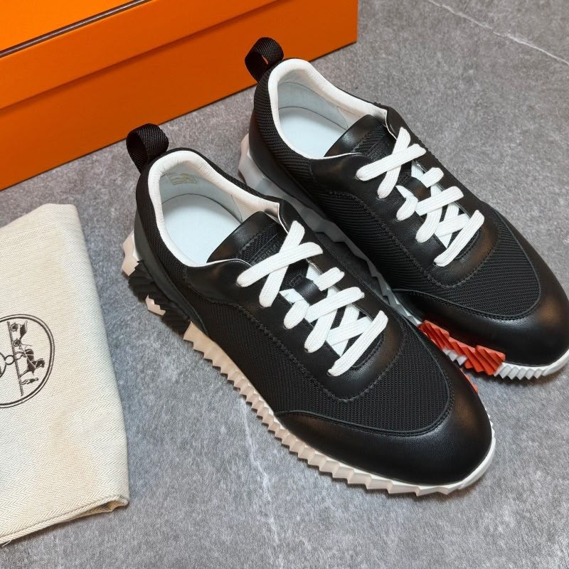 A pair of black leather and mesh sneakers with white laces and white zigzag soles, displayed next to an orange box and a beige dust bag on a gray floor.