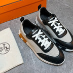 A pair of low-top sneakers with a beige canvas upper, black leather overlays, and white laces, displayed next to an orange shoebox and a branded dust bag on a gray floor.