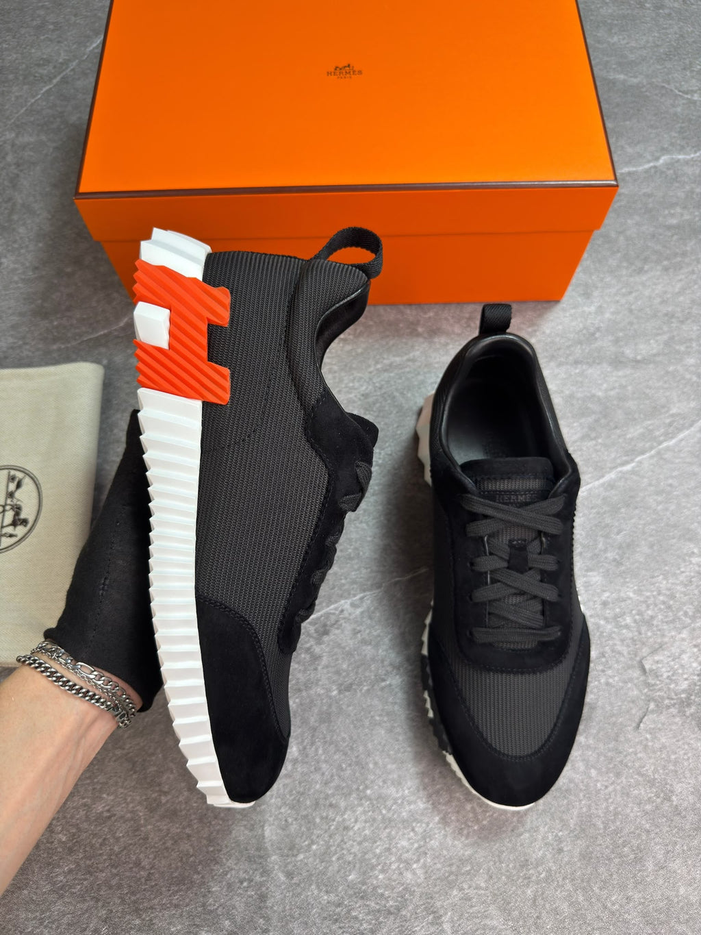 A pair of black athletic sneakers with a distinctive orange and white H-shaped sole, displayed next to an orange Hermès shoebox and a beige dust bag on a gray floor.