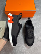 A pair of black athletic sneakers with a distinctive orange and white H-shaped sole, displayed next to an orange Hermès shoebox and a beige dust bag on a gray floor.