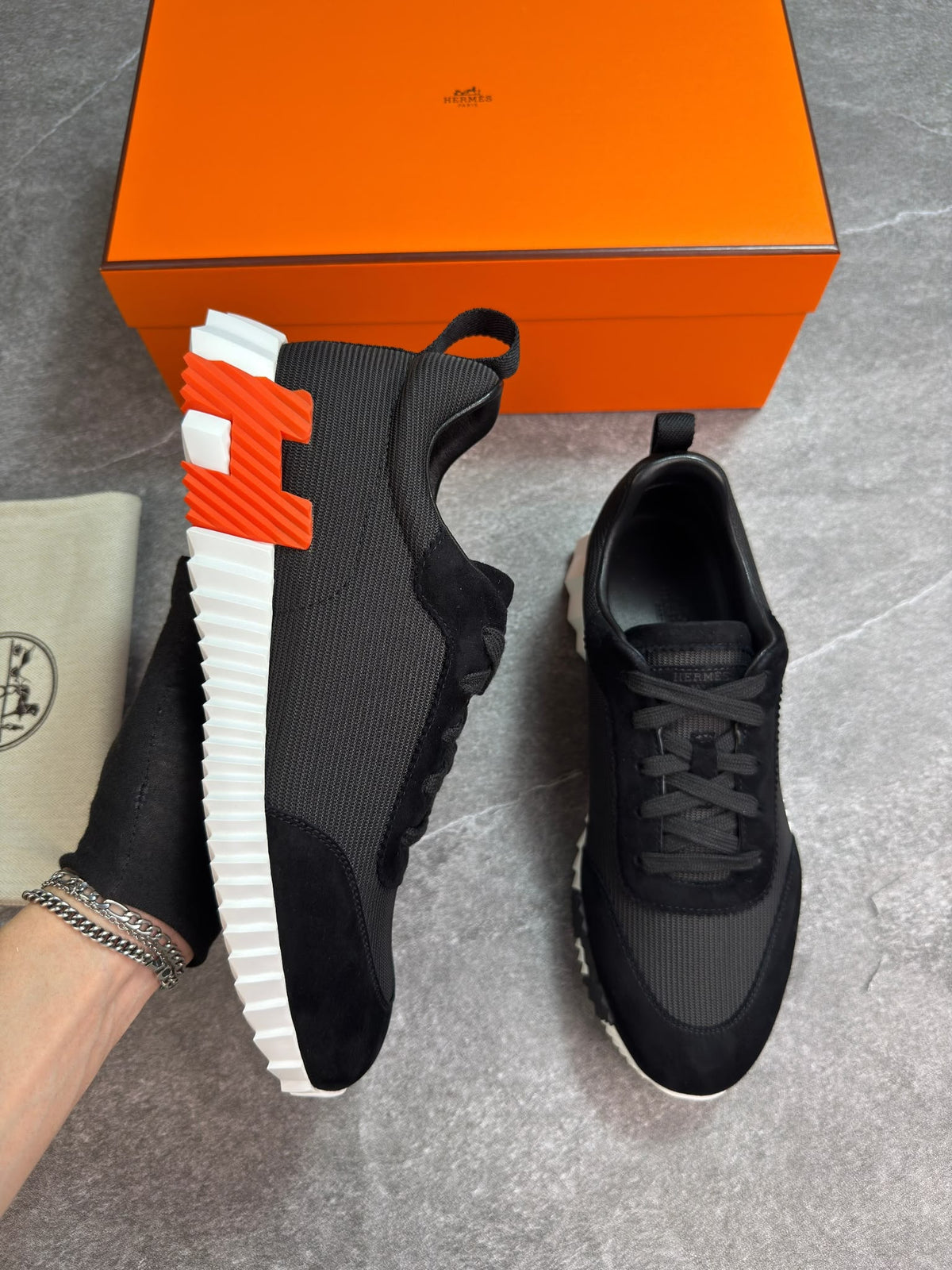 A pair of black athletic sneakers with a distinctive orange and white H-shaped sole, displayed next to an orange Hermès shoebox and a beige dust bag on a gray floor.