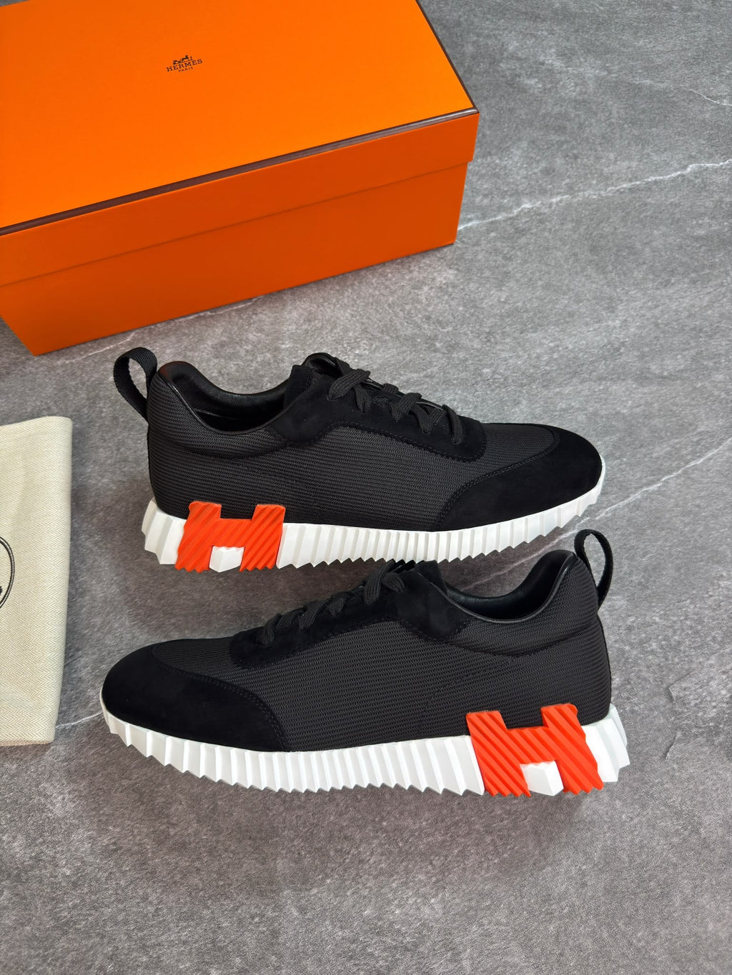 A pair of black low-top sneakers with a bold white ridged sole and a large orange-and-white H logo on the heel, displayed on a gray stone surface with an orange shoe box and dust bag nearby.