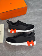 A pair of black low-top sneakers with a bold white ridged sole and a large orange-and-white H logo on the heel, displayed on a gray stone surface with an orange shoe box and dust bag nearby.