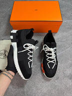 A pair of black knit sneakers with chunky white soles and white laces, displayed next to an orange Hermès box and a branded dust bag on a gray floor.