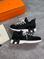 A pair of black low-top sneakers with chunky white ribbed soles and white laces, displayed on a gray stone surface next to an orange shoebox and a dust bag.