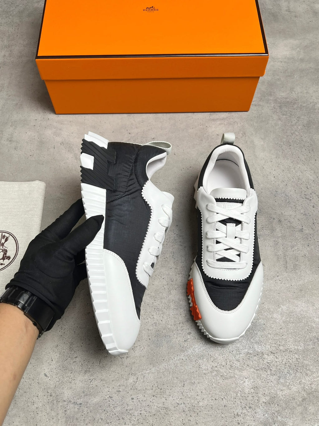 A pair of black and white low-top sneakers displayed on a gray concrete floor, with one shoe held by a gloved hand and the other standing upright. An orange box and a dust bag are visible in the background.