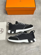A pair of black and white low-top sneakers displayed on a gray concrete surface, with an orange Hermès shoebox and a white dust bag with the Hermès logo visible in the background.