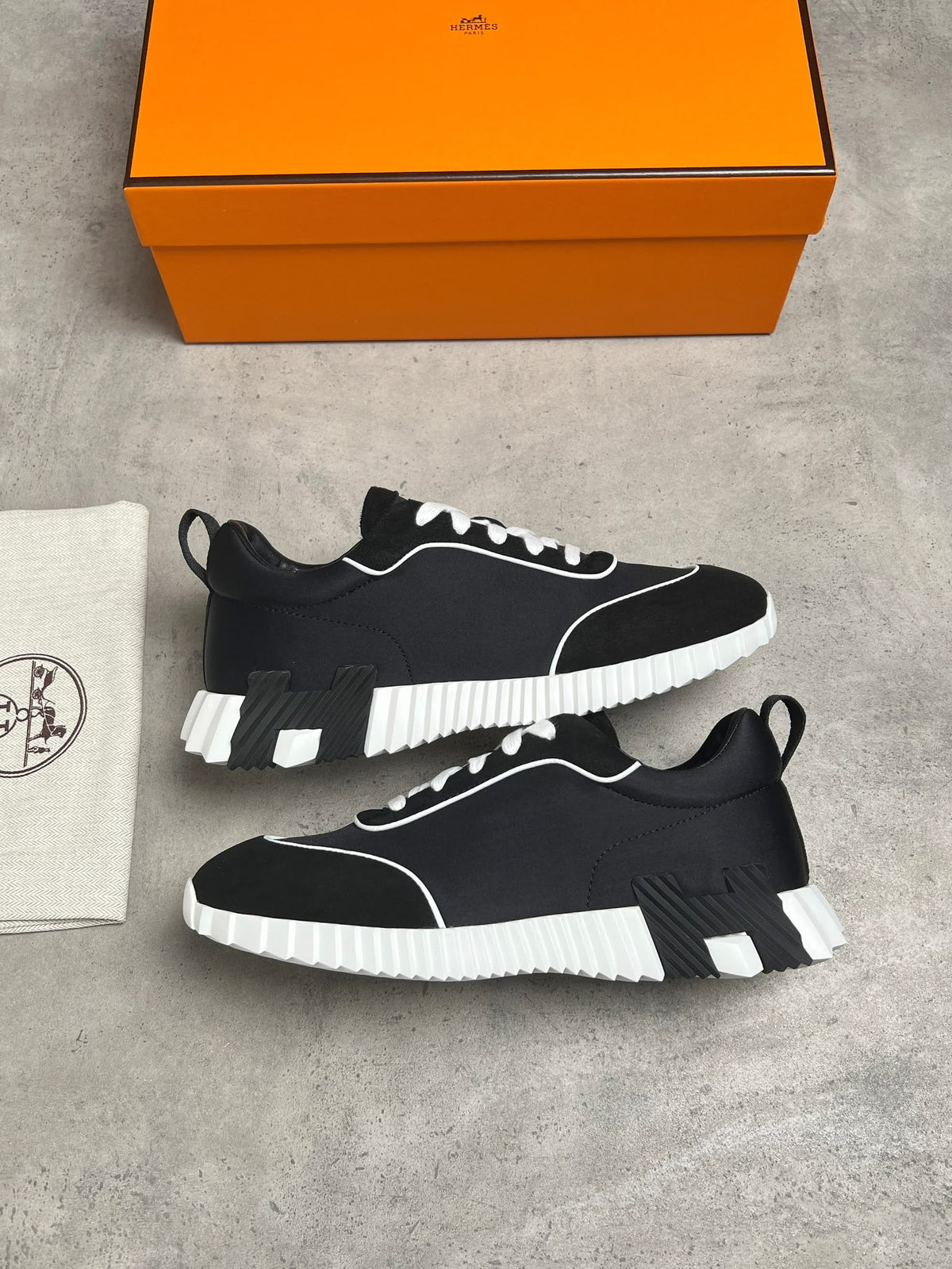 A pair of black and white low-top sneakers displayed on a gray concrete surface, with an orange Hermès shoebox and a white dust bag with the Hermès logo visible in the background.