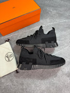 A pair of black low-top sneakers displayed on a gray stone surface, with an orange shoebox and a cream dust bag with a circular logo beside them.