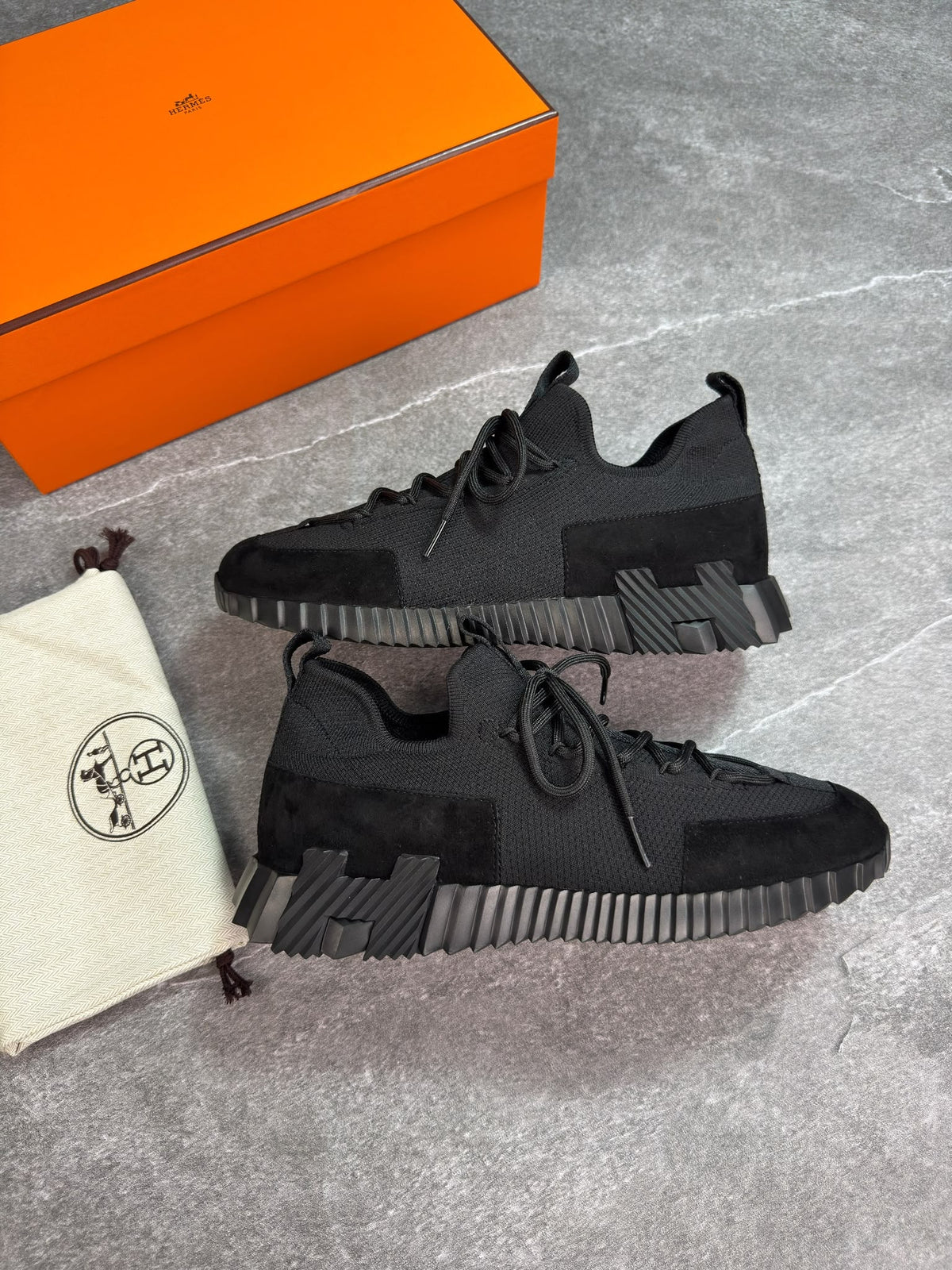 A pair of black low-top sneakers displayed on a gray stone surface, with an orange shoebox and a cream dust bag with a circular logo beside them.