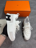 A pair of white low-top sneakers with chunky white soles and black accent details, shown with an orange Hermès shoebox on a gray floor. One shoe is held by a hand wearing a black glove.