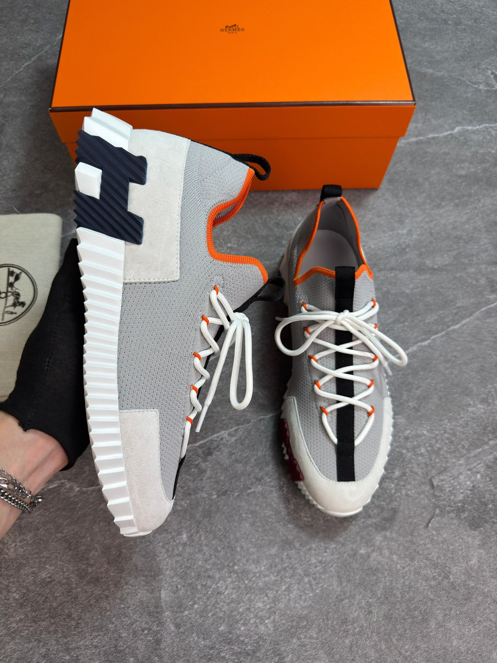 A pair of gray and white sneakers with orange accents and a large black 'H' detail on the side, displayed next to an orange Hermès box on a gray floor.
