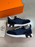 A pair of navy blue and white low-top sneakers with black accents, displayed next to an orange Hermès box and a branded dust bag on a neutral gray surface.