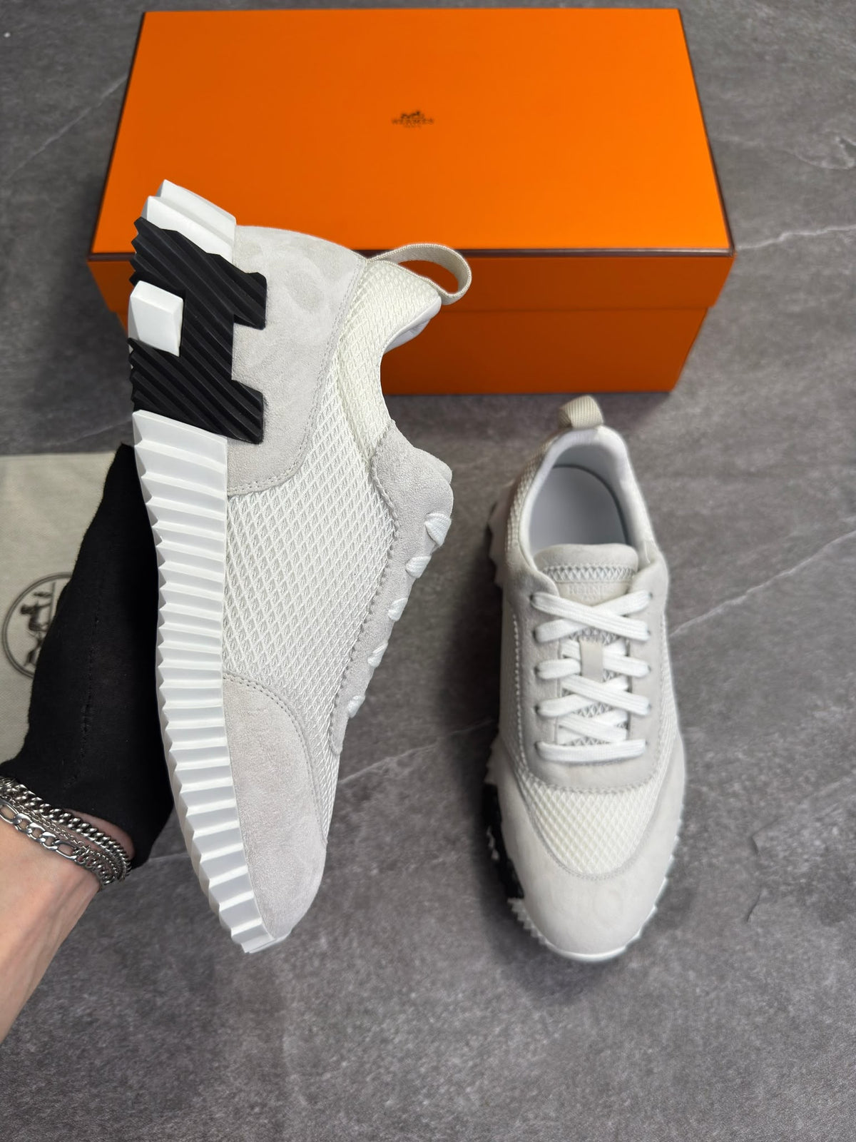 A pair of light gray and white knit sneakers, with one held by a hand wearing a black glove and the other resting on a gray floor. An orange Hermès shoebox is visible in the background.