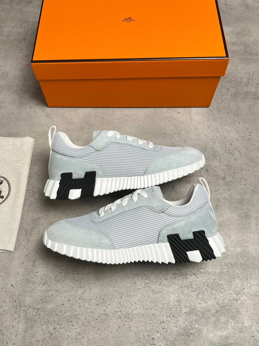 A pair of light gray and white knit sneakers with black H-shaped soles, displayed on a gray concrete surface next to an orange Hermès box and a dust bag.