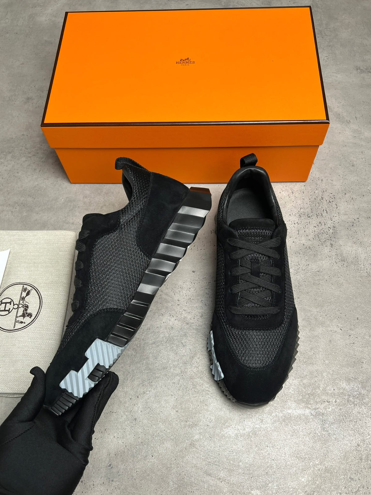 Black sneakers with a silver stripe on a gray surface next to an orange box.