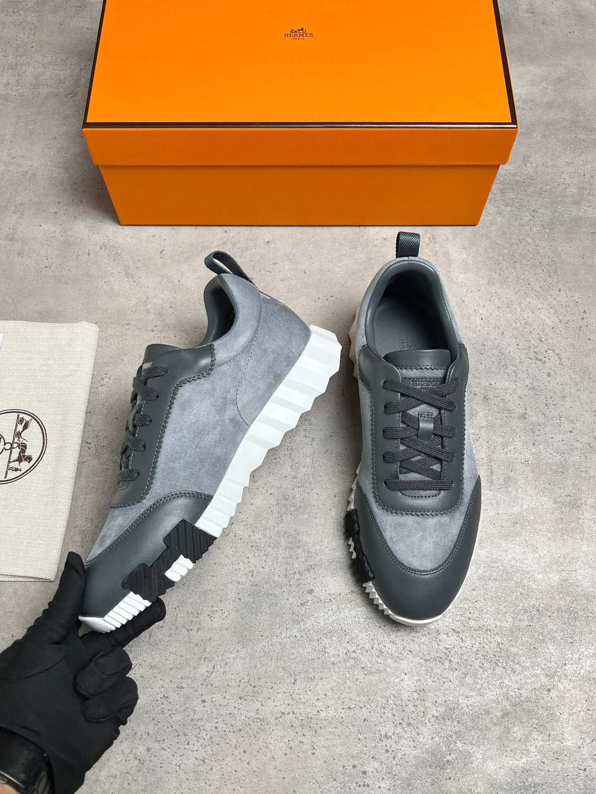 A pair of gray and black low-top sneakers with chunky white platform soles, displayed on a gray concrete floor next to an orange Hermès box and a branded dust bag. A black gloved hand is holding one shoe.