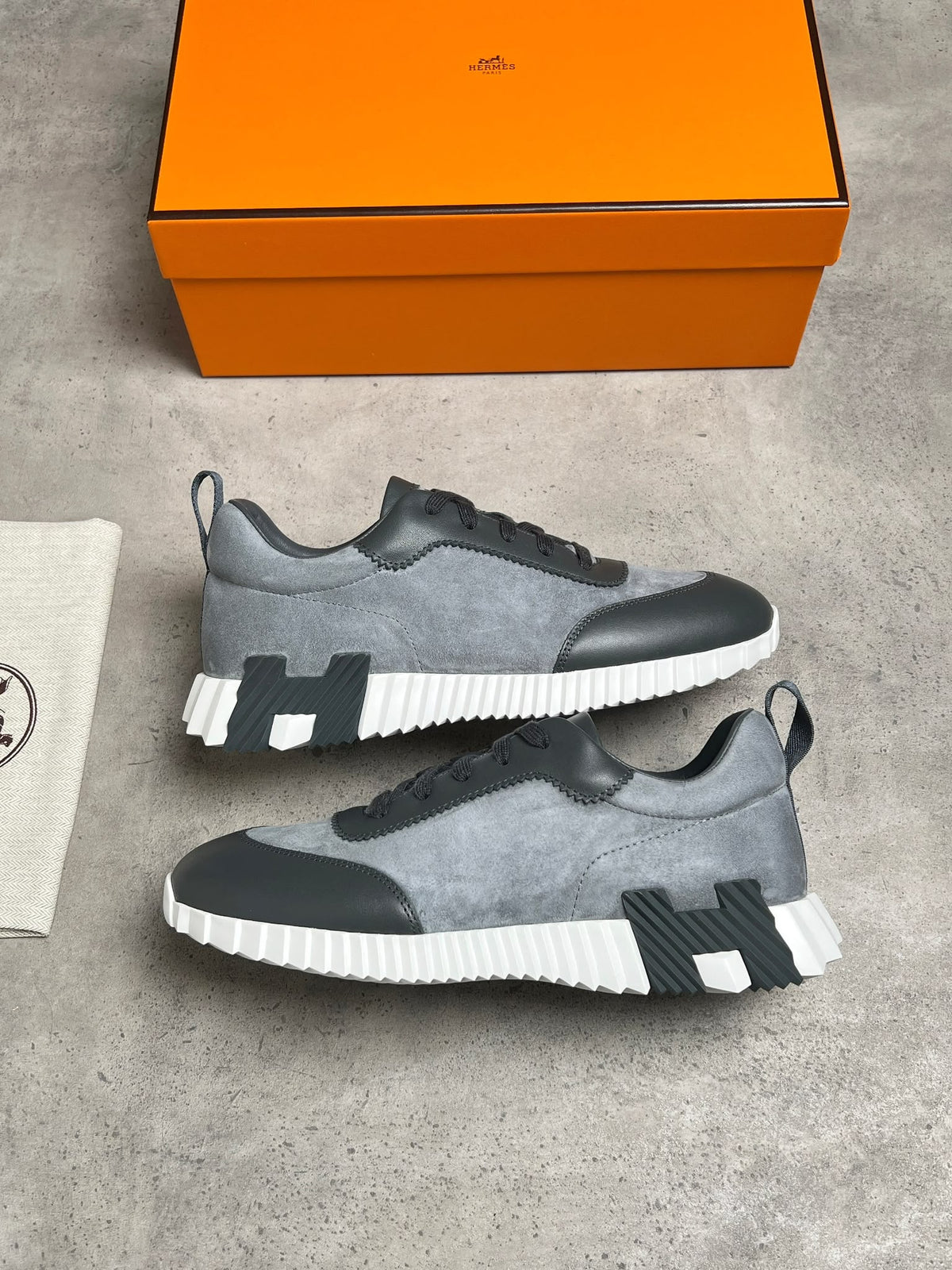 A pair of gray and black low-top sneakers with white sculpted soles and a large black H logo on the side, displayed on a gray concrete surface with an orange shoebox and dust bag in the background.