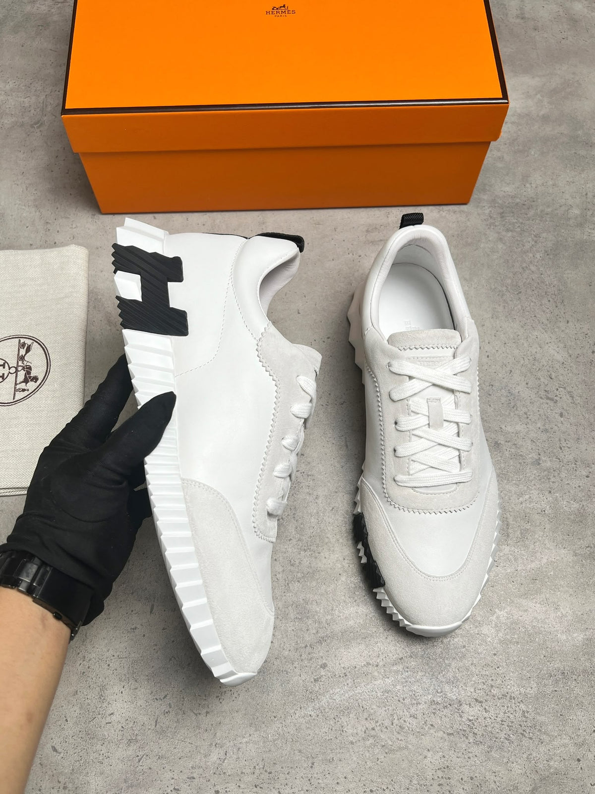 A pair of white Hermès sneakers with black accents, shown with a black-gloved hand holding one shoe, an orange Hermès box, and a branded dust bag on a gray concrete surface.