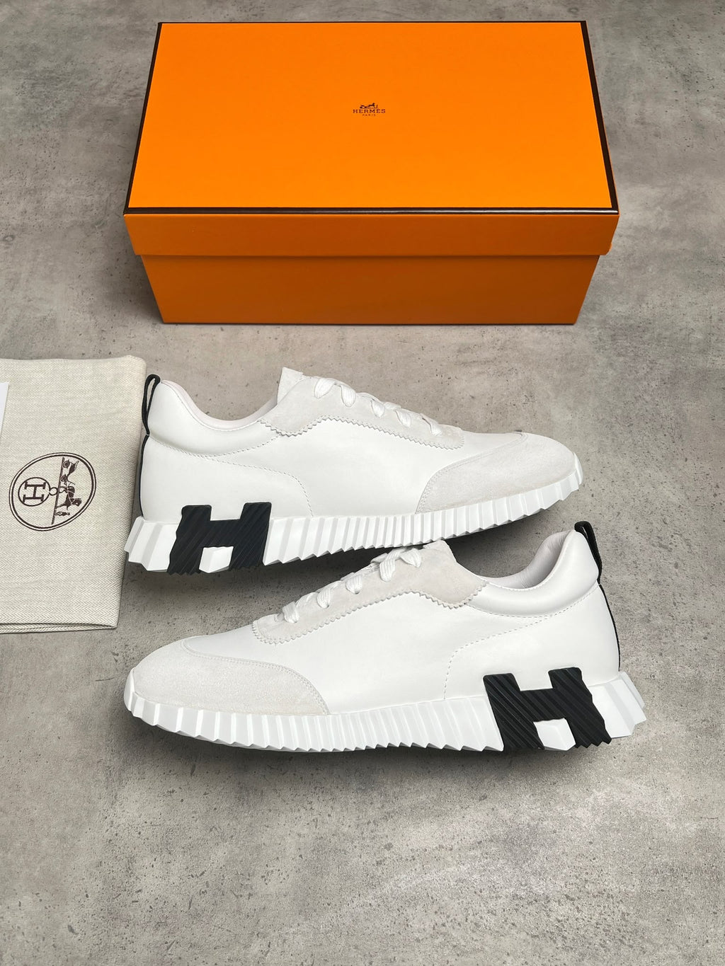 A pair of white low-top sneakers with black H-shaped side accents, displayed on a gray concrete surface with an orange Hermès shoebox and a branded dust bag.