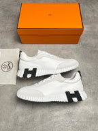 A pair of white low-top sneakers with black H-shaped side accents, displayed on a gray concrete surface with an orange Hermès shoebox and a branded dust bag.