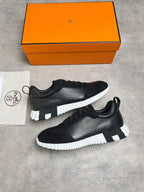 A pair of black leather sneakers with chunky white sculpted soles, displayed next to their signature orange Hermès shoebox and branded dust bag on a gray concrete surface.
