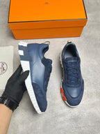 A pair of navy blue sneakers with white soles, shown with an orange Hermès box and a black-gloved hand holding one shoe. The Hermès logo is visible on the box and dust bag.