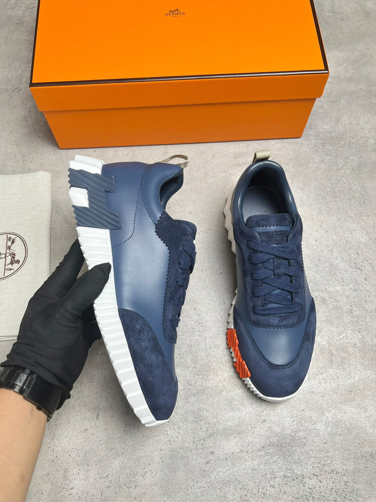 A pair of navy blue sneakers with white soles, shown with an orange Hermès box and a black-gloved hand holding one shoe. The Hermès logo is visible on the box and dust bag.