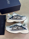 A pair of Dior low-top sneakers in white, gray, and navy blue, displayed next to their navy blue Dior shoebox and a navy blue dust bag on a neutral beige background.