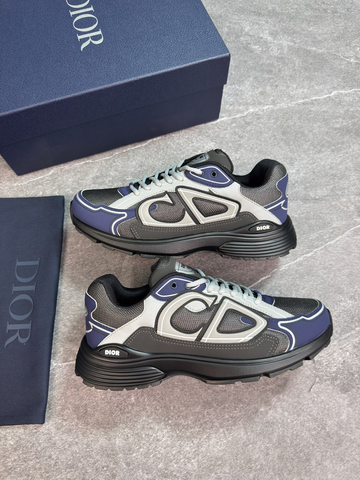 A pair of Dior sneakers in navy blue, gray, and black, displayed next to their navy blue Dior shoebox on a gray stone-textured background.