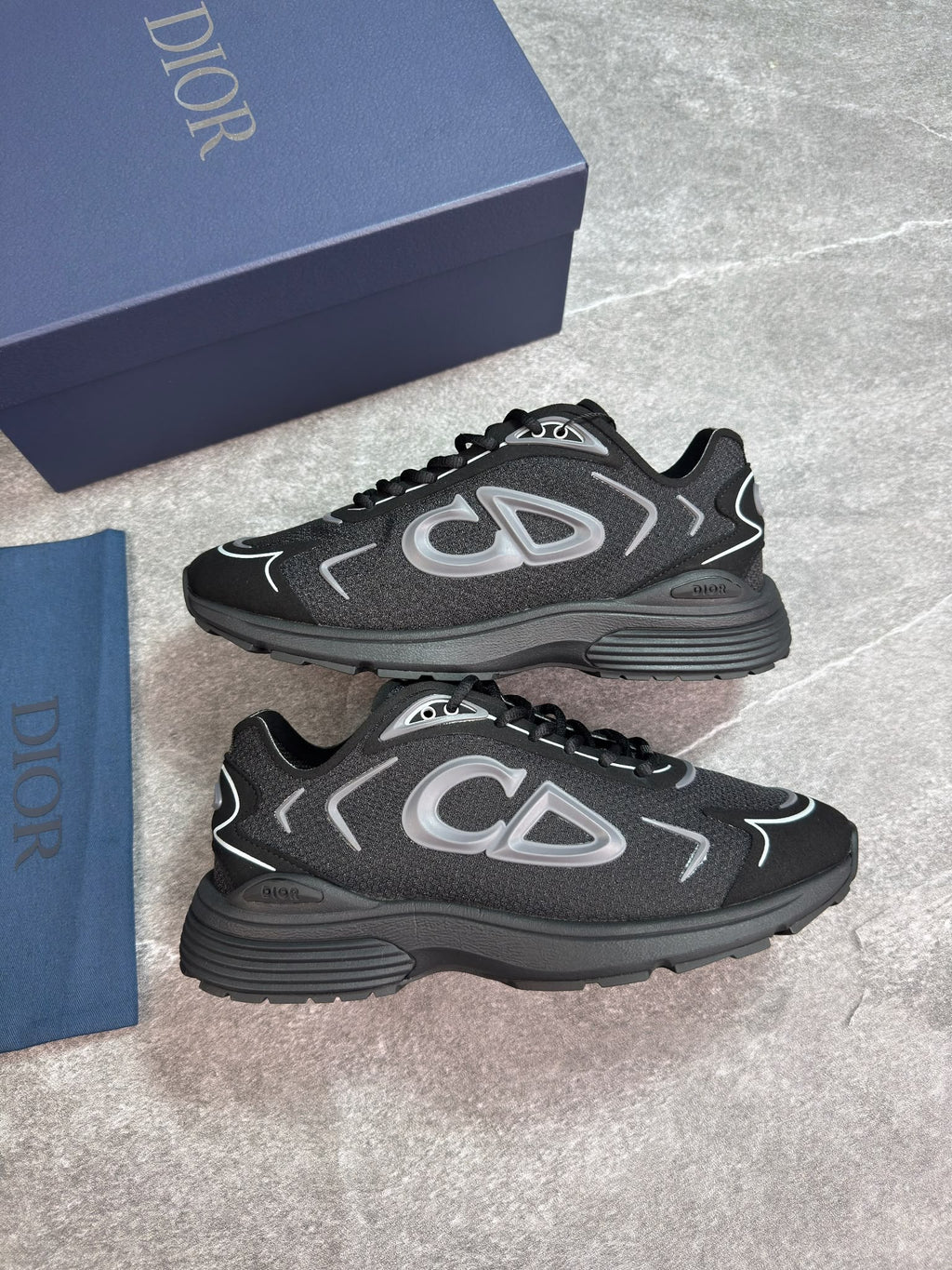 A pair of Dior low-top sneakers in black and dark gray with silver accents, displayed next to their navy blue Dior shoebox and dust bag on a gray stone-textured background.
