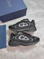 A pair of Dior low-top sneakers in black and dark gray with silver accents, displayed next to their navy blue Dior shoebox and dust bag on a gray stone-textured background.