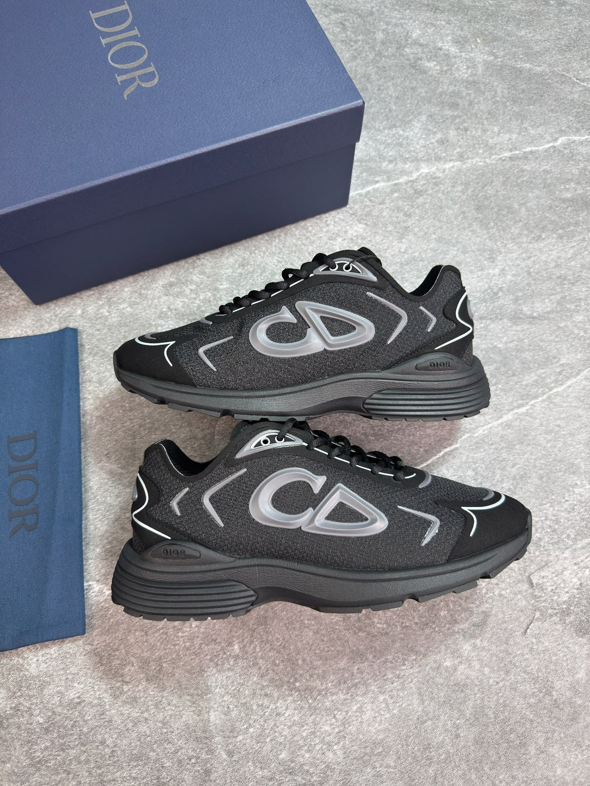 A pair of Dior low-top sneakers in black and dark gray with silver accents, displayed next to their navy blue Dior shoebox and dust bag on a gray stone-textured background.
