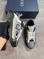 A pair of Dior sneaker sneakers in black, gray, and off-white, with one shoe held by a gloved hand and the other standing on a gray floor, with a navy blue Dior shoebox behind them.