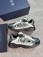 A pair of Dior low-top sneakers in black, gray, and off-white, displayed next to their navy blue Dior box and dust bag on a gray stone-textured surface.