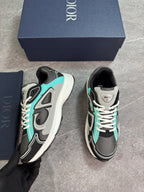 A pair of Dior sneakers in black, gray, and turquoise, shown with their navy blue Dior shoebox and dust bag on a gray floor. A hand wearing a black glove is holding one shoe.
