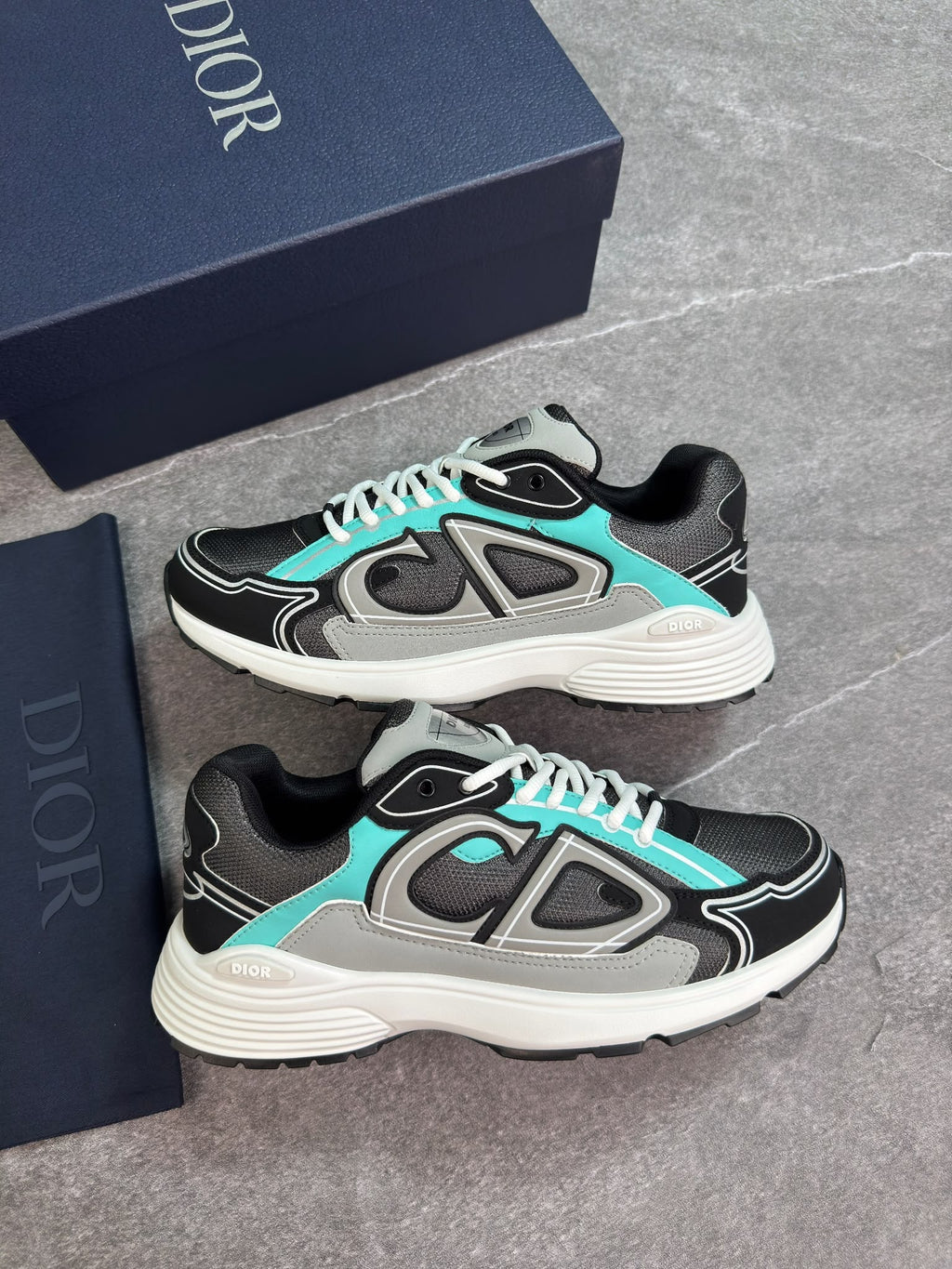 A pair of Dior sneakers in black, gray, and turquoise, displayed next to their navy blue Dior packaging boxes on a gray stone-textured surface.