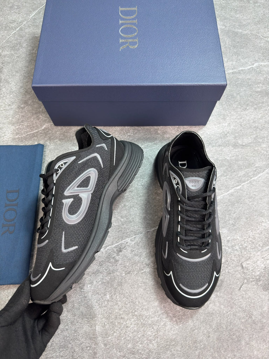 A pair of Dior sneakers in black and gray with silver accents, displayed next to their blue Dior box and dust bag on a gray marble floor.