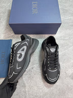 A pair of Dior sneakers in black and gray with silver accents, displayed next to their blue Dior box and dust bag on a gray marble floor.