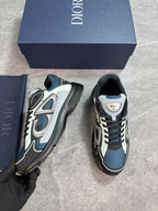 A pair of Dior sneaker sneakers in navy blue, black, and white, shown with their navy blue Dior shoebox and a navy blue Dior dust bag. A hand wearing a black glove holds one of the sneakers next to the other which is placed on a gray stone floor.