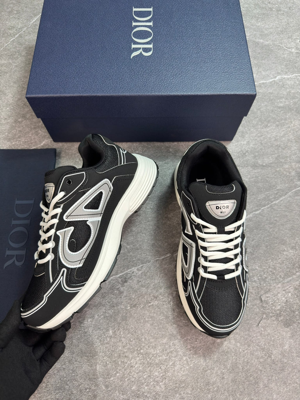 A pair of Dior low-top sneakers in black and white with silver accents, displayed next to their navy blue Dior shoebox on a gray tiled floor.