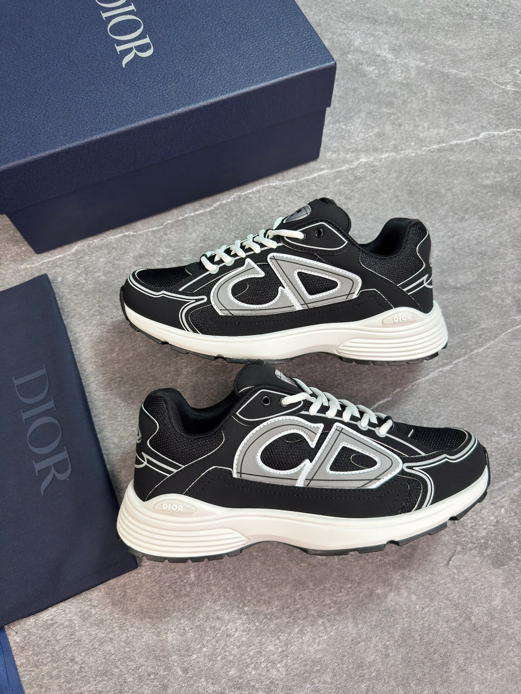 A pair of Dior sneakers in black and silver, displayed next to their navy blue Dior shoebox on a gray stone-textured background.