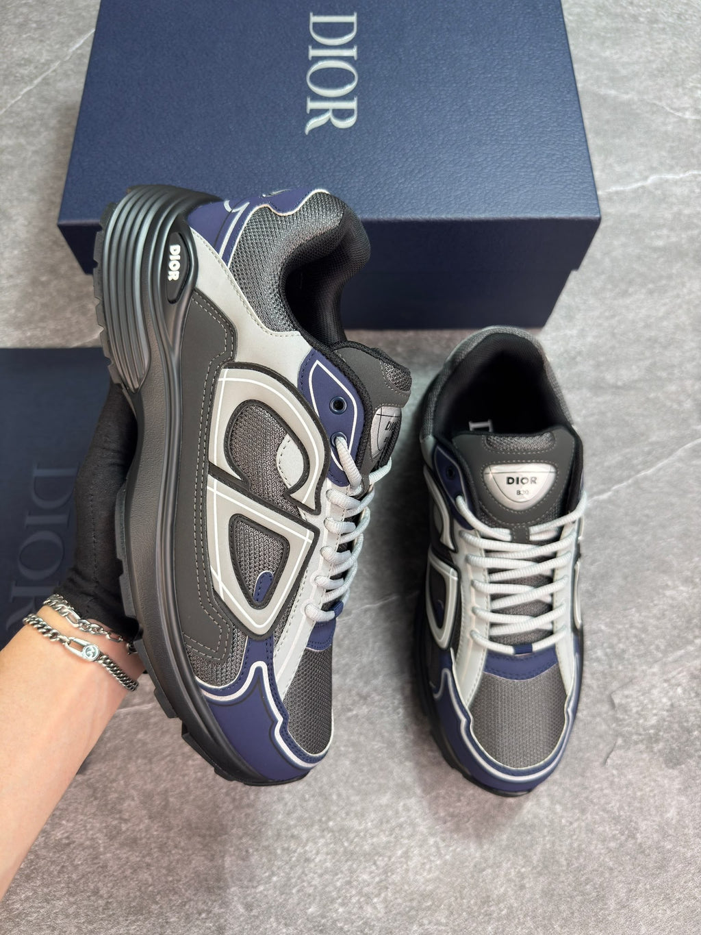 A pair of Dior sneakers in navy blue, gray, and white, with one shoe held by a hand wearing a black glove and the other resting on a gray floor. A navy blue Dior shoebox is visible in the background.