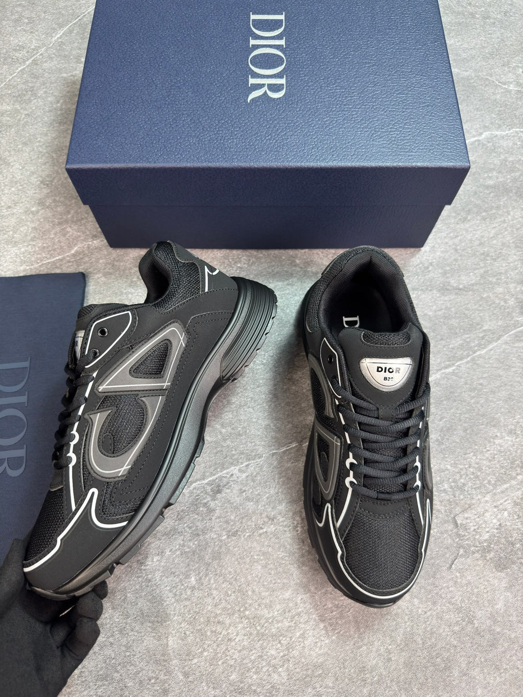 A pair of Dior low-top sneakers in black with gray and white accents, shown next to their navy blue Dior shoebox on a gray tiled floor.
