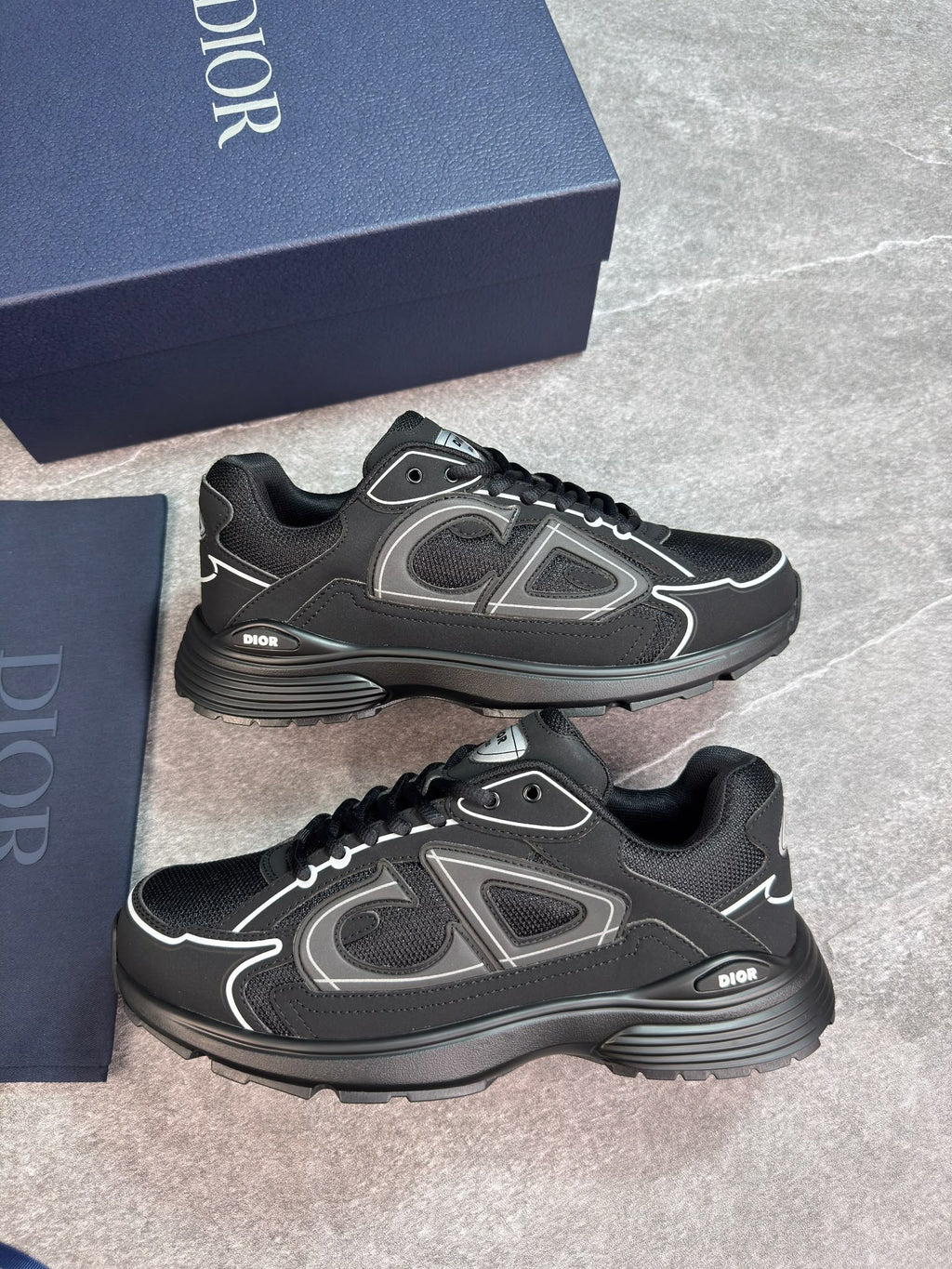 A pair of Dior low-top sneakers in black with gray and white accents, displayed next to their navy blue Dior shoebox on a gray stone floor.